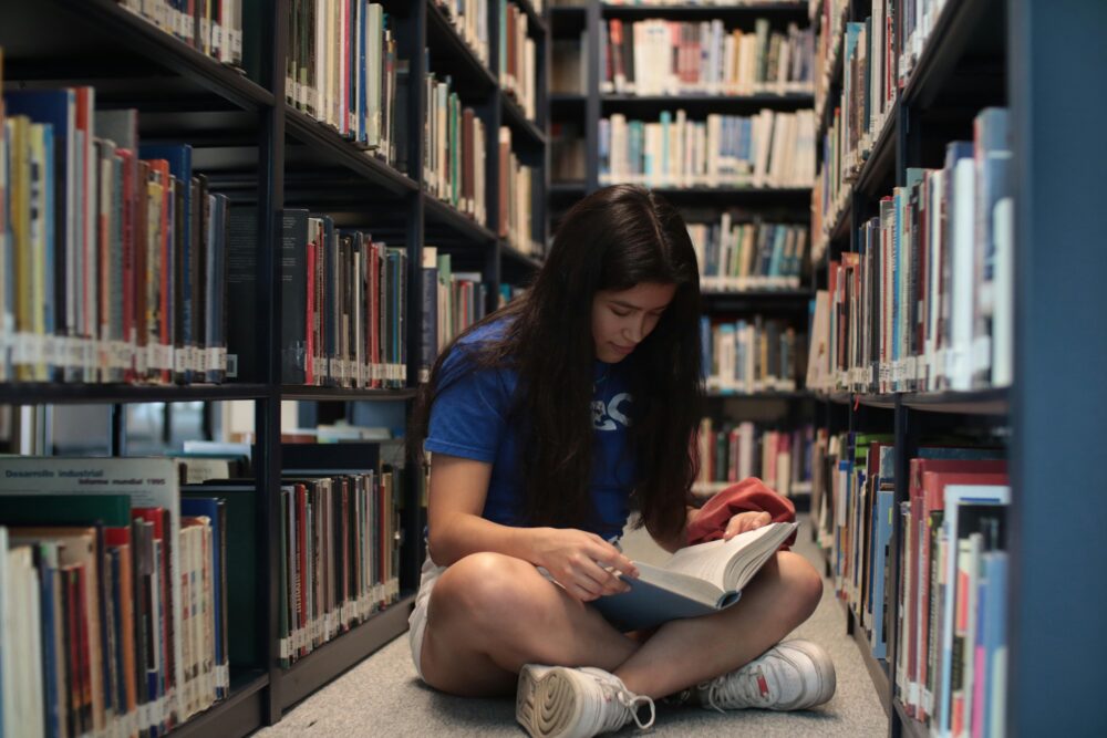 Teenage girl sitting on the floor of a library, reading a book. (Photo by Ludovic Delot via Pexels)
