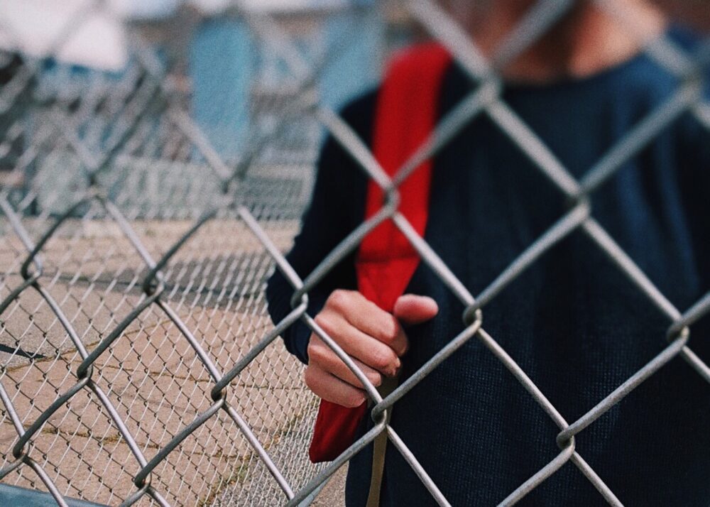 Midsection of a student wearing a backpack and standing behind a wire fence. (Image by WOKANDAPIX on Pixabay)