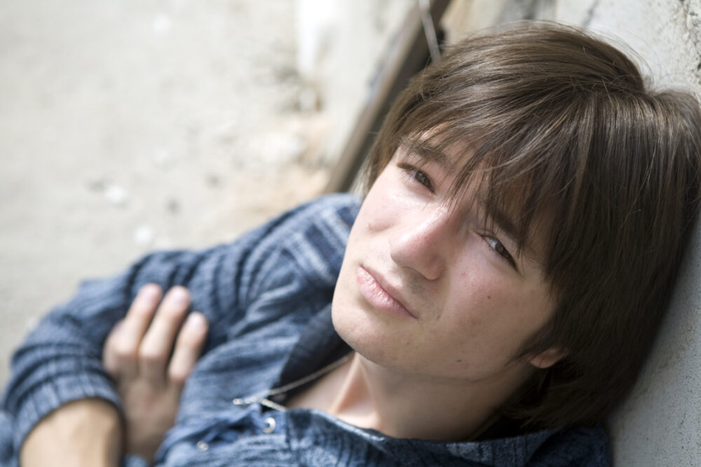Teenage boy leans against a wall, arms crossed.