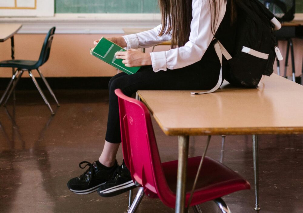 Girl sitting on a desk in a classroom, reading a book. (Photo by RDNE Stock project from Pexels)
