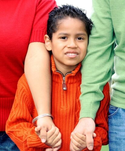 A young boy in an orange sweater stands between two adults, holding each of their hands.