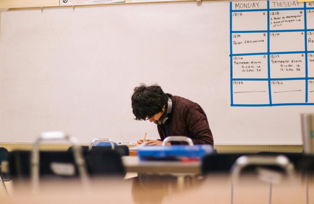 Older male student writing on a table at the front of a classroom. (Photo by Jeswin Thomas from Pexels)