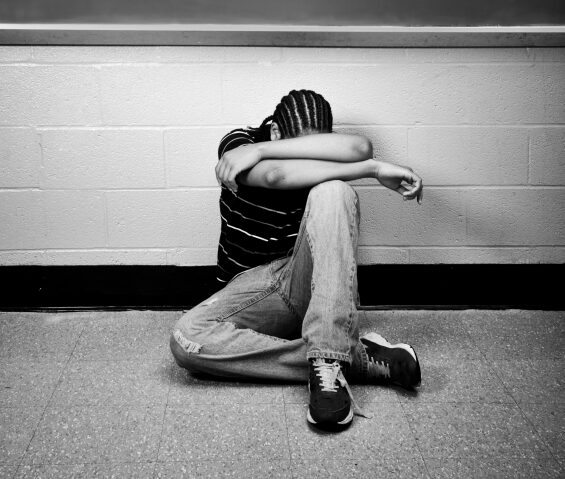 Male teenager hides his head in his arms as he sits on the floor of a classroom.