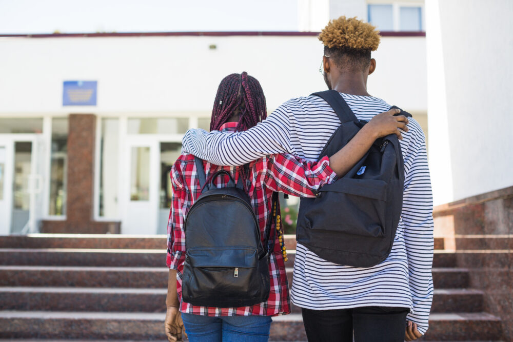 Two teenagers walk up the steps of a school, arms around one another. (Image by Freepik)