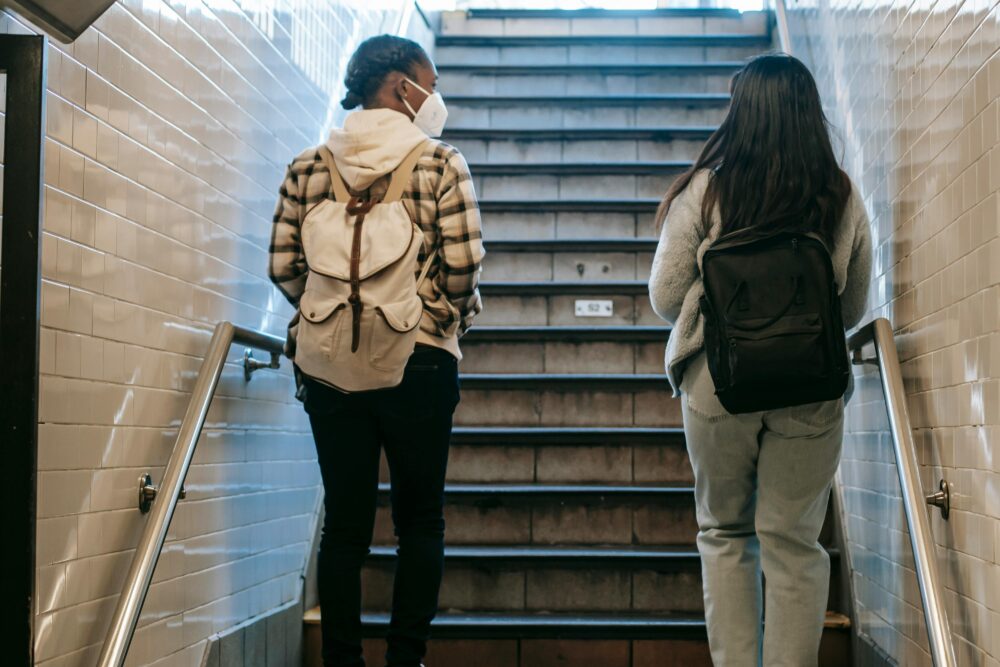 Two teenage girls with backpacks, one wearing a face mask, walk up a flight of stairs in a subway station. (Photo by Charlotte May via Pexels)