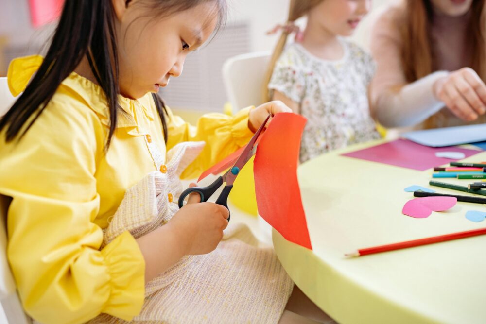 Preschooler in a yellow dress cuts a sheet of red construction paper.