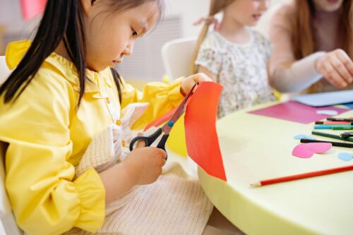 Preschooler in a yellow dress cuts a sheet of red construction paper.