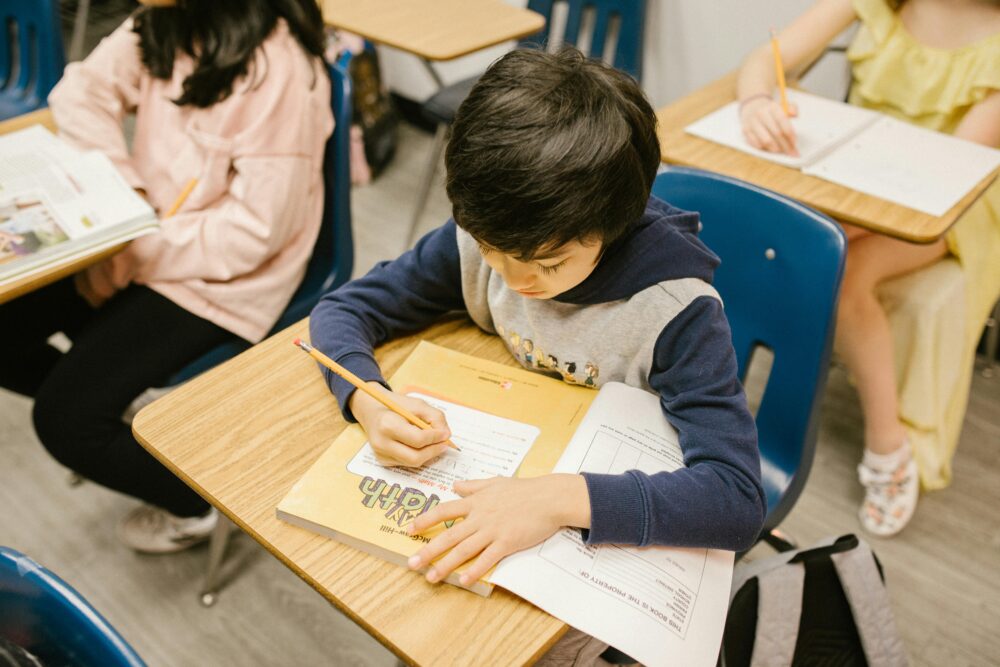 Boy sitting at a desk in a classroom, writing in a workbook. (Photo by RDNE Stock project via Pexels)