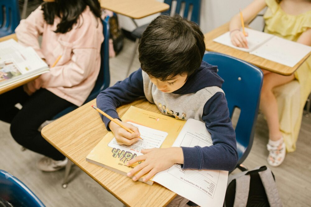 Boy sitting at a desk in a classroom, writing in a workbook.