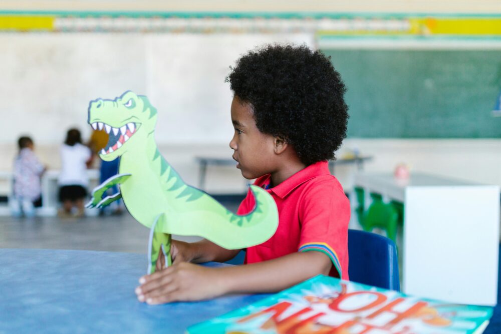 Side view of a Black preschool boy in a classroom, holding a large dinosaur cutout. (Photo by RDNE Stock project via Pexels)