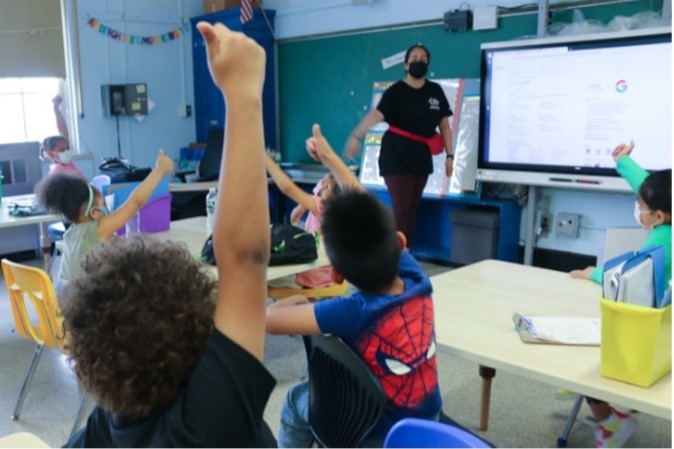 Young students raise their hands and give a thumbs up to their teacher during a summer program. New York City will change the admissions method for its sprawling summer enrichment program.