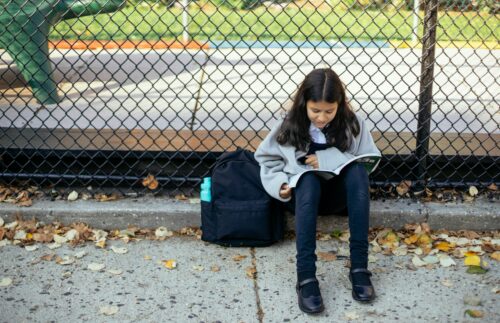 A girl sits with her backpack, looking at a textbook, outside a schoolyard. (Photo by Mary Taylor via Pexels)