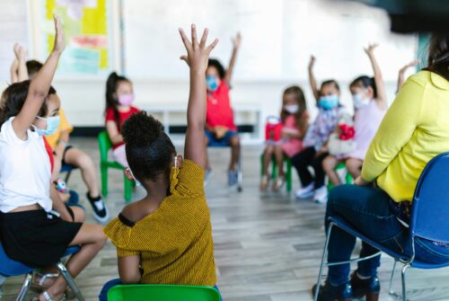 Young students sitting in a circle, several raising their hands. (Photo by RDNE Stock project via Pexels)