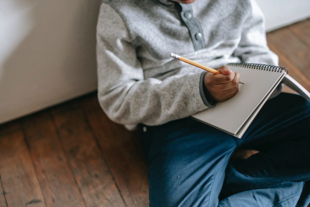 Midsection of a student sitting on the floor, writing in a notebook with a pencil. (Photo by Katerina Holmes from Pexels)