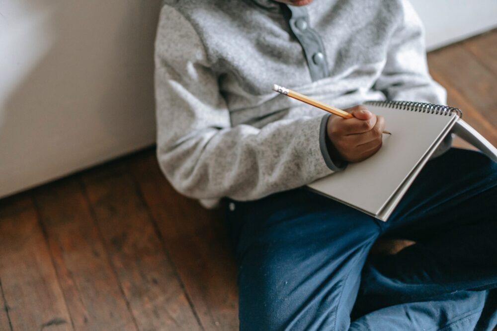 Midsection of a student sitting on the floor, writing in a notebook with a pencil.