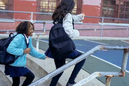 Two girls in school uniforms run up the stairs in a school yard.