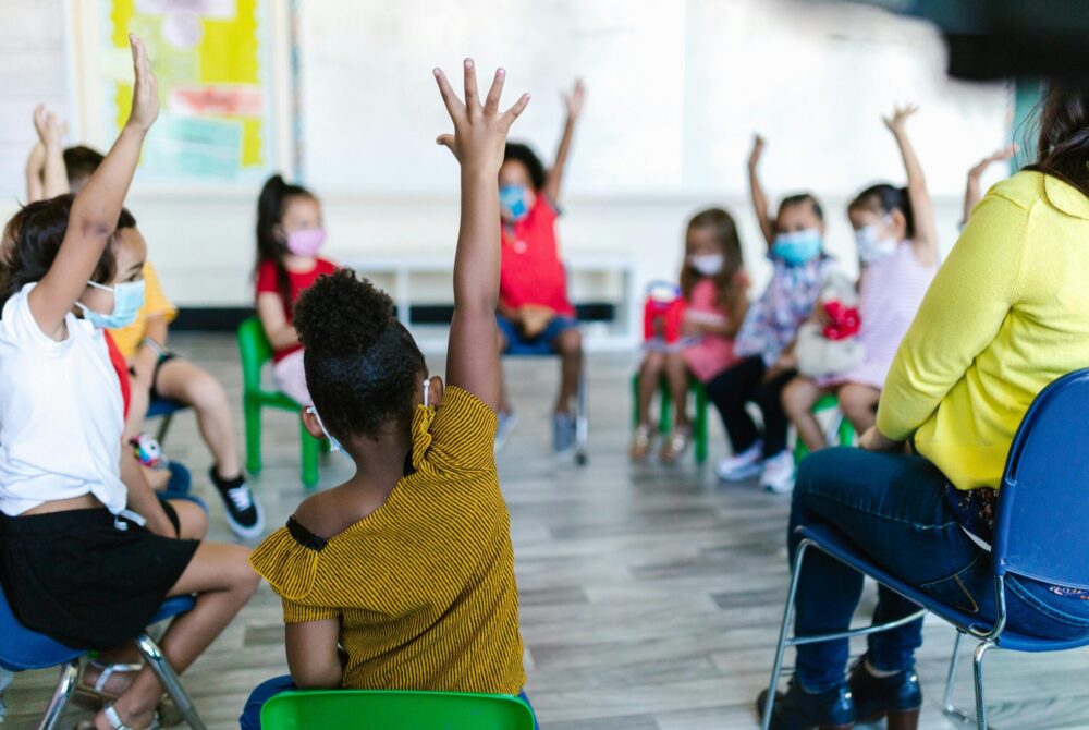 Young students sitting in a circle, several raising their hands.