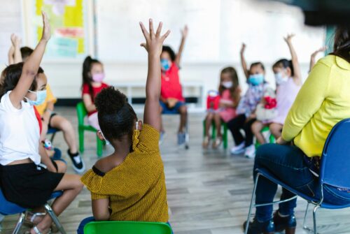 Young students sitting in a circle, several raising their hands.