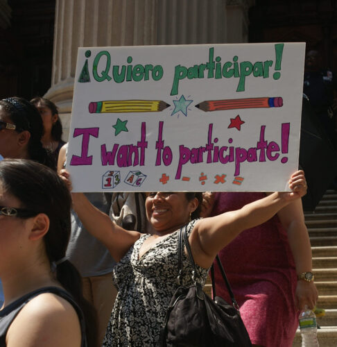 Parent holds a sign at a rally reading 'Quiero participar! I want to participate!'