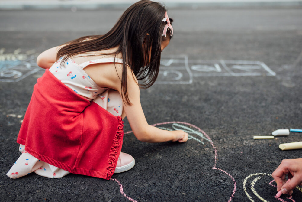 Side view image of little girl drawing with colorful chalks on playground.