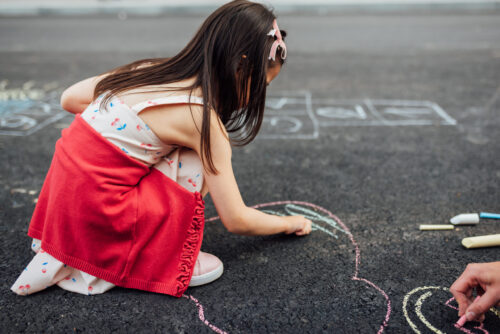 Side view image of little girl drawing with colorful chalks on playground.