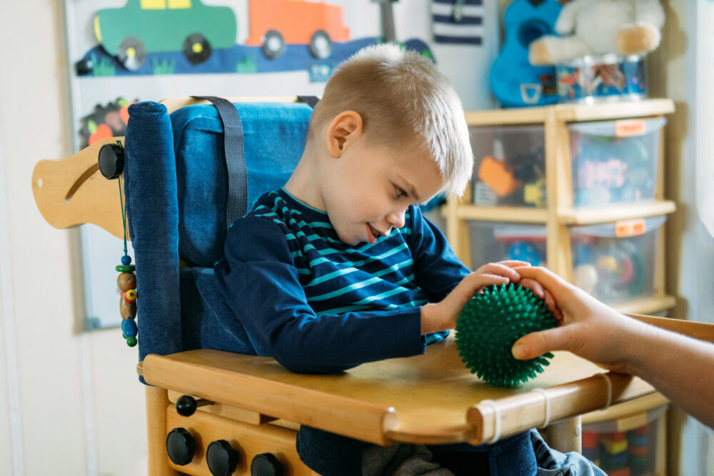 Boy with with cerebral palsy in special chair playing with a textured ball. (Photo by irissca, Adobe Stock)