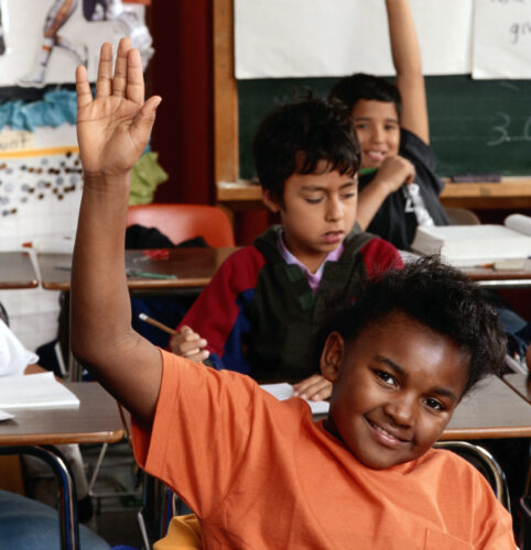 A student in the front row of a classroom raises their hand.