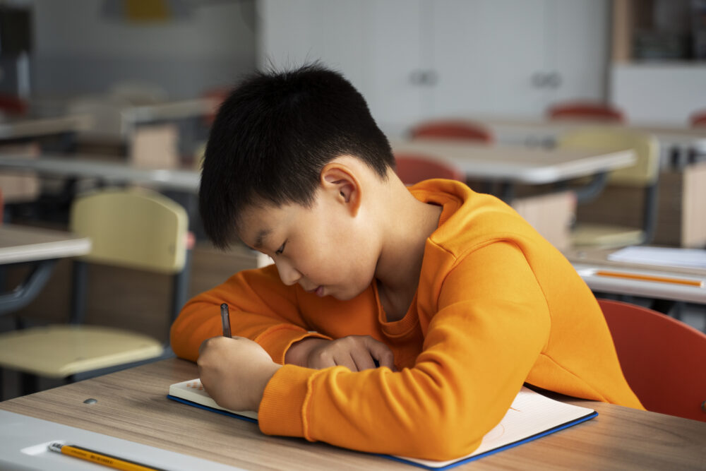 Elementary-school-age Asian boy sitting at a desk, writing in a book. (Image by Freepik)