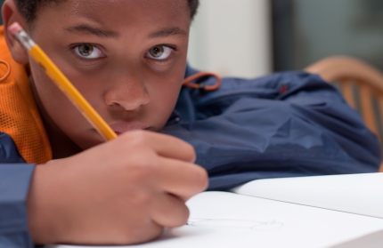 A young boy looks at the camera, holding a pencil and writing in a notebook