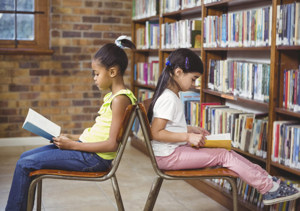 Two young girls sit back-to-back in a library, each reading a book.