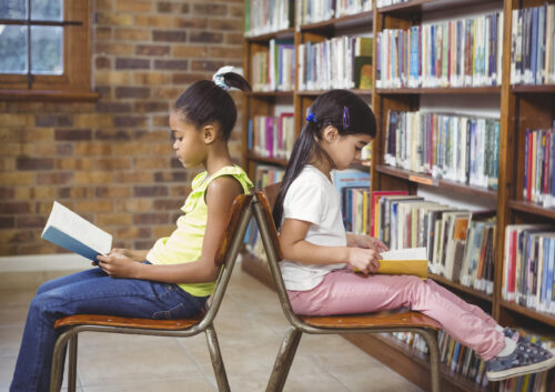 Two young girls sit back-to-back in a library, each reading a book.