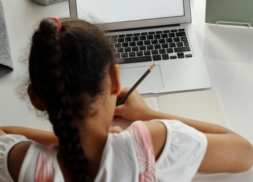 A young girl sits in front of a laptop, taking notes with a pencil. (Photo by August de Richelieu via Pexels)