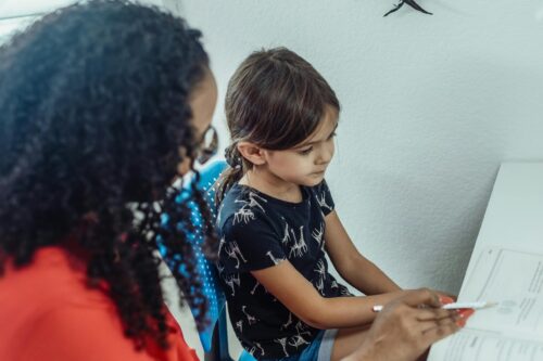 Woman sits next to a young girl, pointing at a workbook.