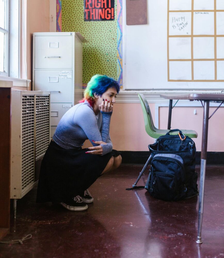 Teen girl with rainbow-colored hair squats in the corner of a classroom, looking at the floor. (Photo by RDNE Stock Project via Pexels)
