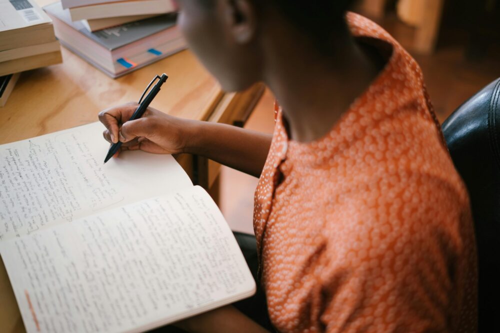 Rear shot of a young woman writing in a notebook; textbooks are piled in the background.
