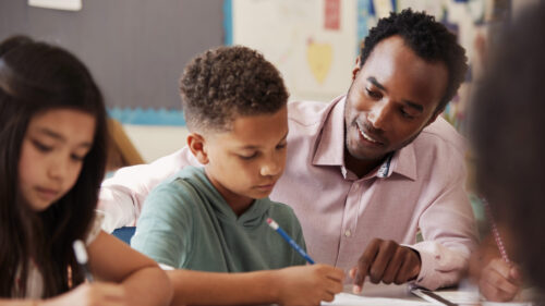 Male teacher working with elementary school boy at his desk