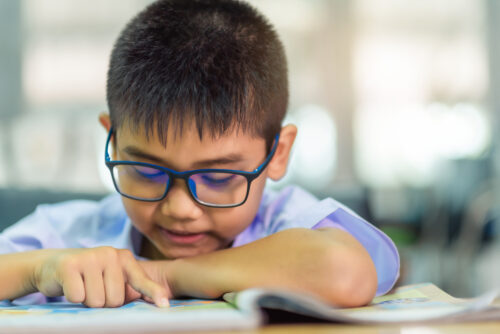 Elementary school boy with glasses reads a book while tracking the text with his finger. (Photo by pop_thailand, Adobe Stock)