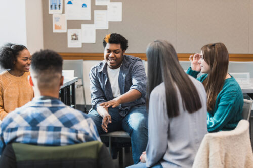 Group of teenagers sit in a circle; a boy gestures as he talks about something with the people in the group. (Photo by SDI Productions, iStock)