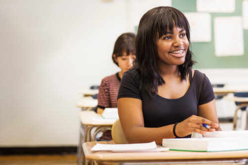 Teenage girl sitting at desk in classroom, smiling at someone off-camera