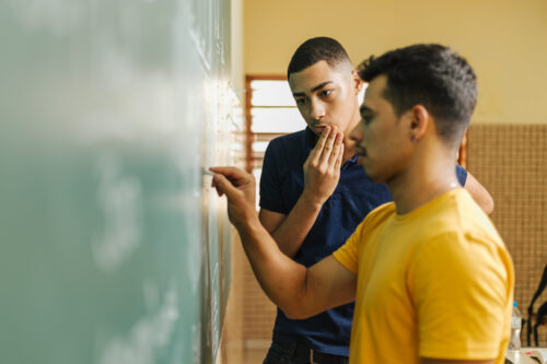 Two Latinx teenage boys at a chalkboard, one writing while the other looks on thoughtfully