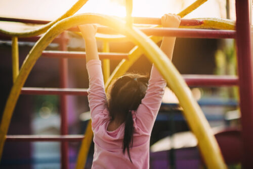 Young girl, viewed from behind, hanging on playground monkey bars.