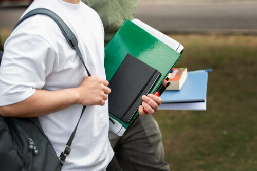 Midsection of two students walking outside, carrying school supplies