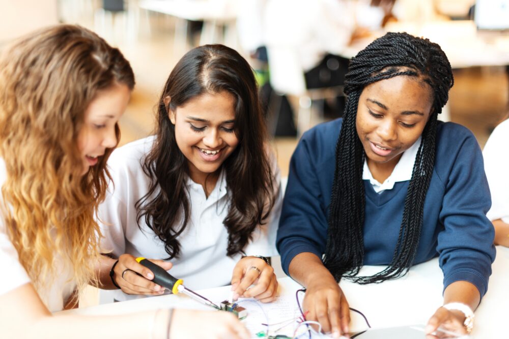 Three young women collaborating on an electronics project, smiling and engaged.