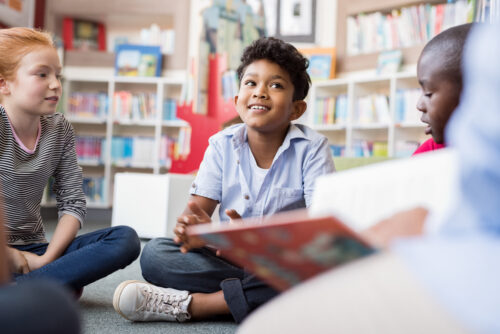 Multiethnic group of elementary schoolers sitting on floor of library, listening a story.