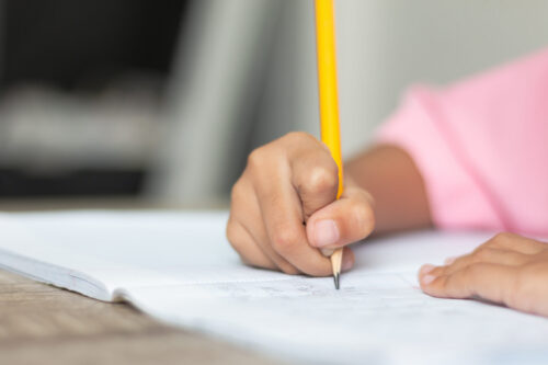 Close up a little girl doing homework. Hand is holding a yellow pencil and writing in a notebook.
