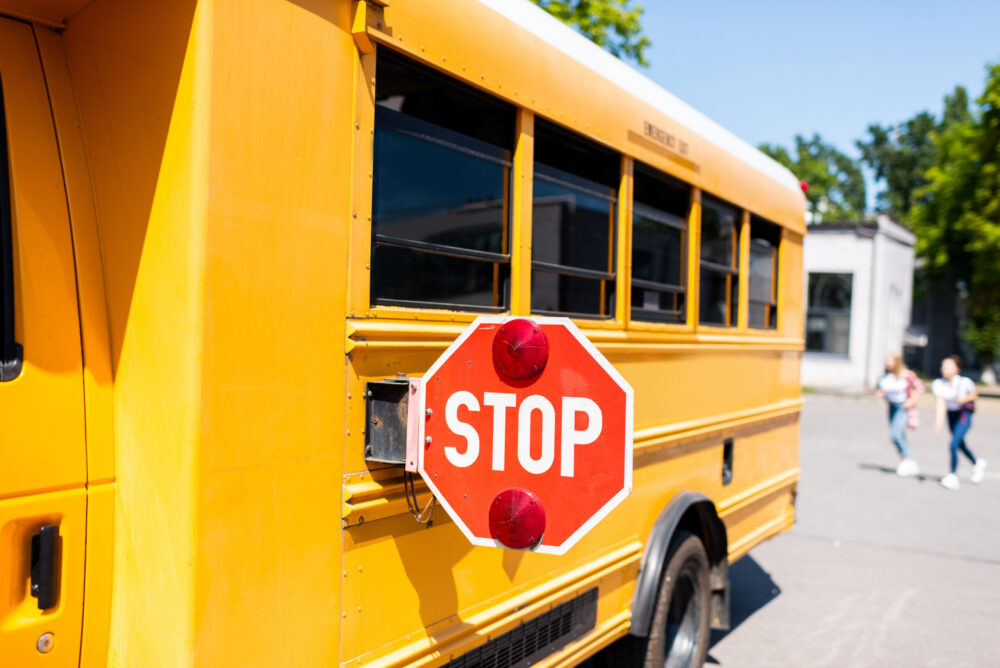 Partial view of parked school bus with stop sign, blurred students running in the background.