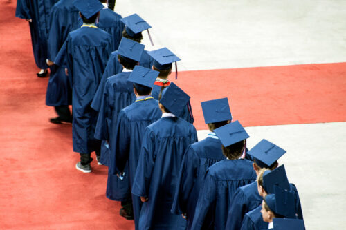 Line of students in blue caps and gowns, viewed from behind.