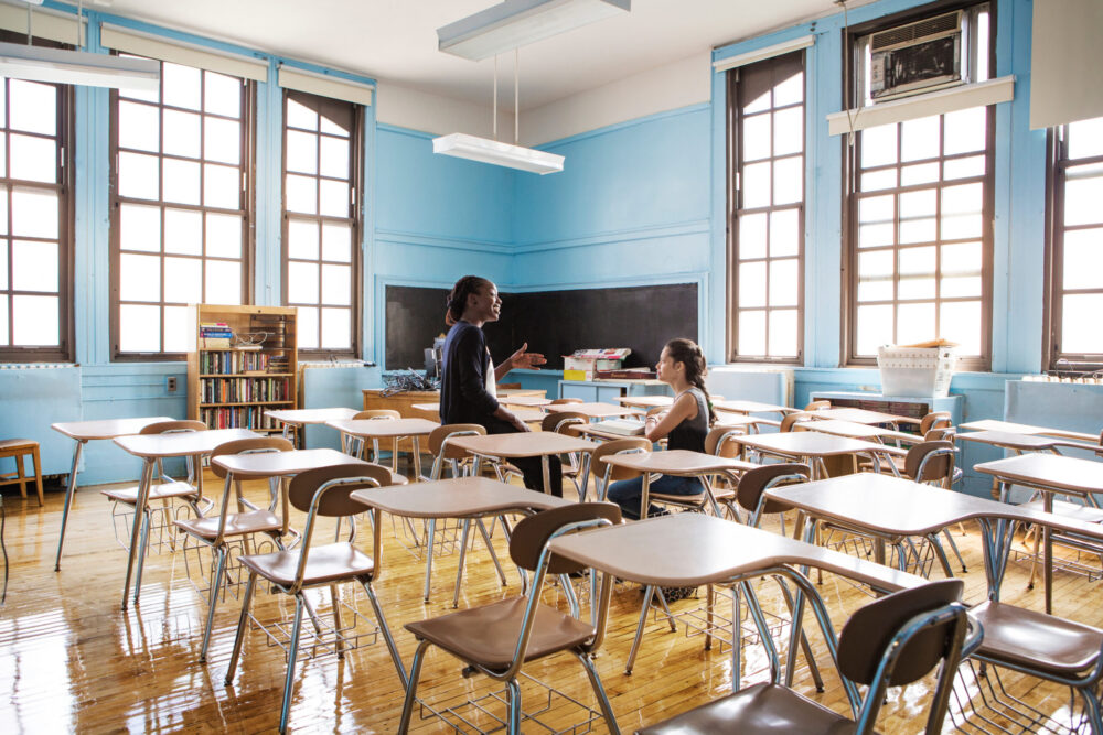 Teenage girl talking with teacher while sitting in classroom.