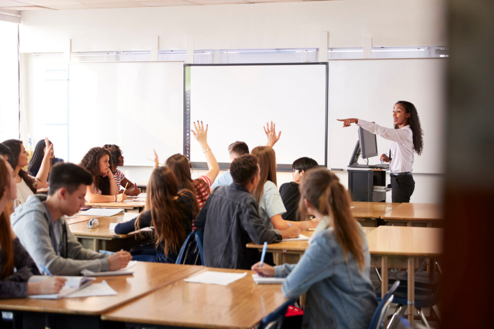Teacher at whiteboard at front of classroom, teaching a class of students