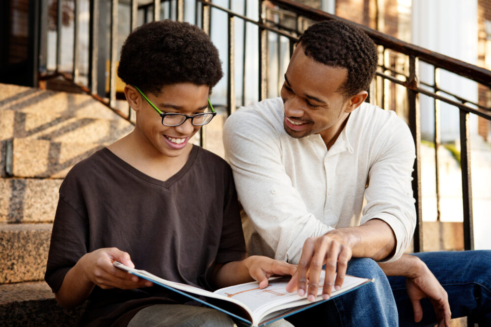 Black boy sitting on steps and reading with an adult. (Photo by Cavan for Adobe, Adobe Stock)